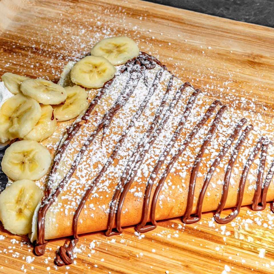 Chunky Bites banana crepe drizzled with chocolate sauce and powdered sugar on a wooden board.