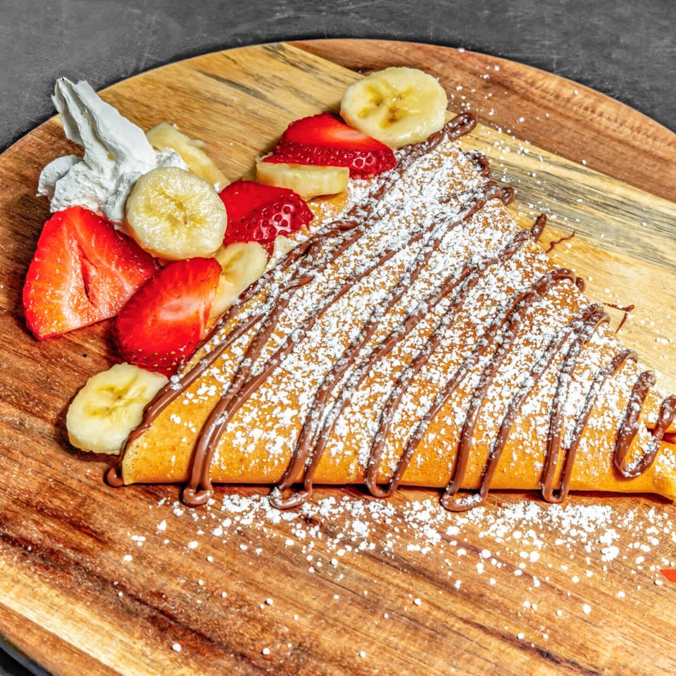 A Chunky Bites crepe drizzled with chocolate and powdered sugar, served with strawberries, bananas, and whipped cream.